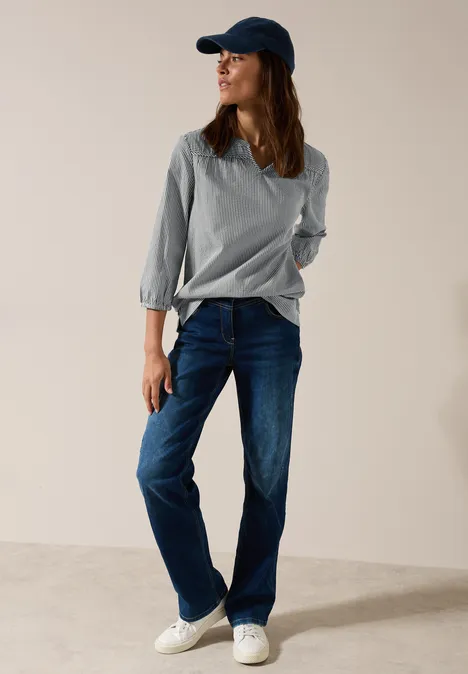 Frau mit Kappe, gestreiftem Oberteil, blauer Jeans und weißen Sneakers steht lässig.